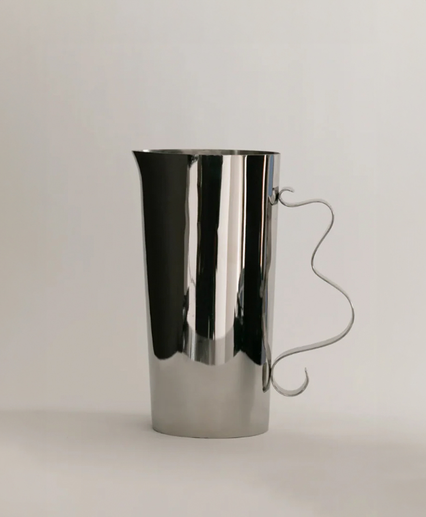 A shiny, silver metal pitcher with a tall, cylindrical shape and a uniquely curvy wire handle, set against a plain light background.