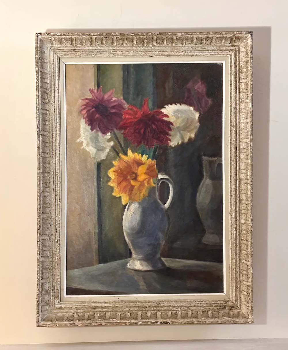 A framed painting of a vase with colorful flowers, including red, yellow, white, and purple blooms, placed on a table with a dark background and a partial reflection of the vase.