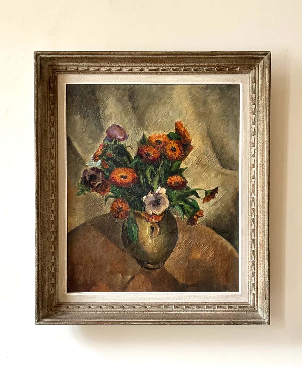 A framed painting of orange and purple flowers in a green vase, set on a brown table with a draped beige cloth in the background, hangs on a cream-colored wall.