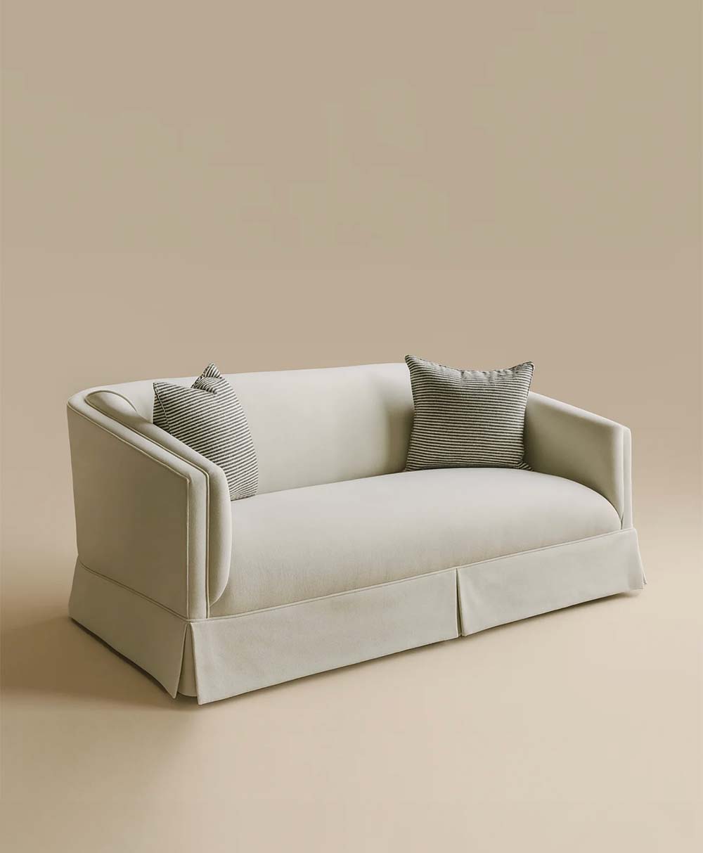 A modern cream-colored sofa with a skirted base, rounded arms, and two striped gray throw pillows, set against a matching neutral background.