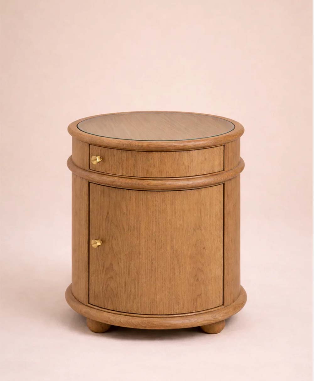 A round wooden side table with a single drawer and a cabinet door, featuring small gold knobs. The table has a smooth finish and stands on four short round feet against a plain background.