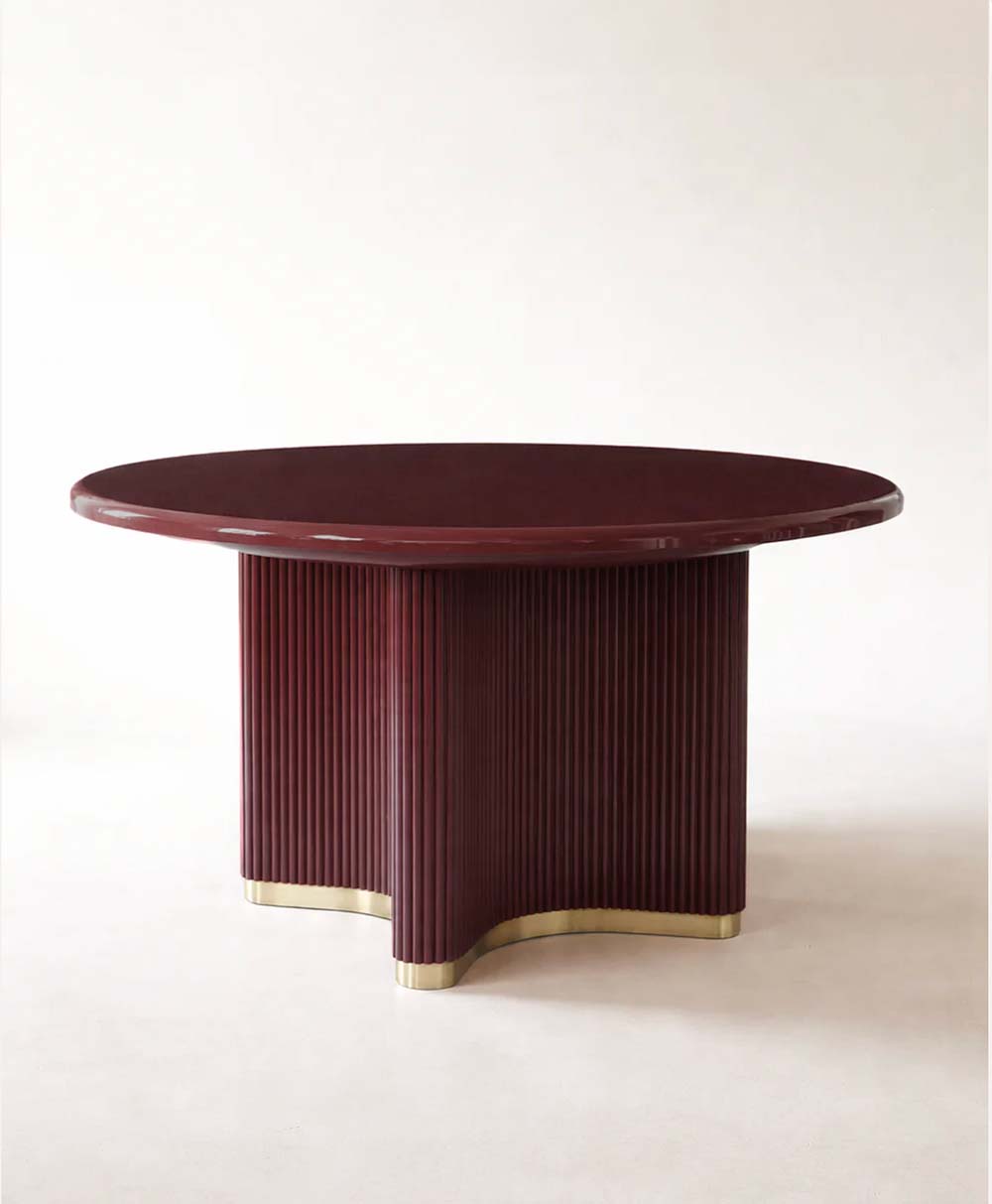 A round, deep red wooden table with a fluted, four-legged pedestal base and gold trim at the bottom, set against a plain, light background.