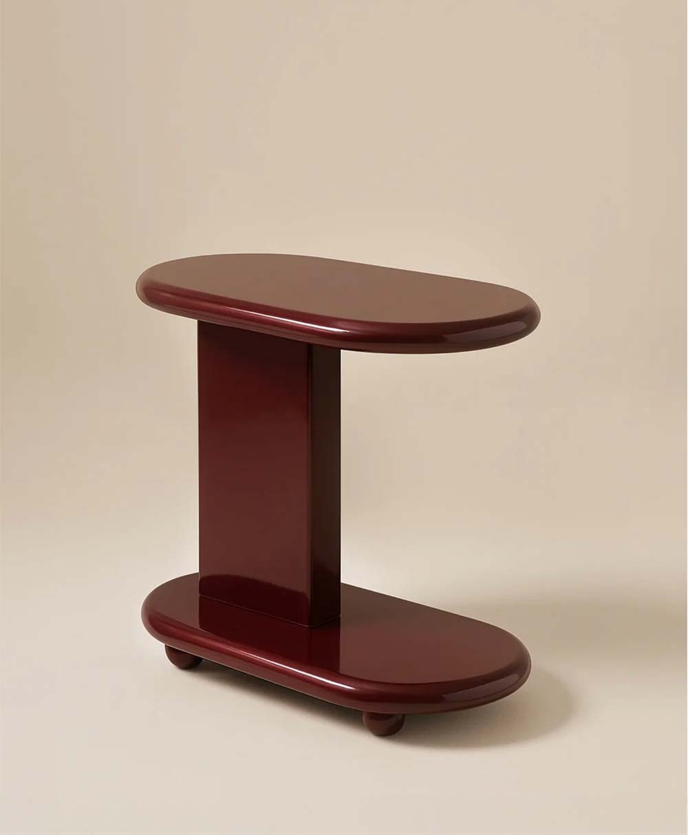 A small, glossy, dark red side table with rounded edges, featuring an oval top and base connected by a central vertical support, set against a plain beige background.