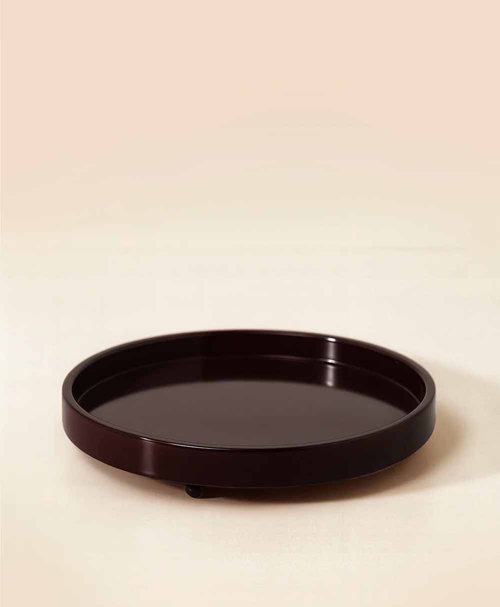 A round, dark brown, glossy serving tray with raised edges is placed on a light, neutral-colored surface against a pale beige background.