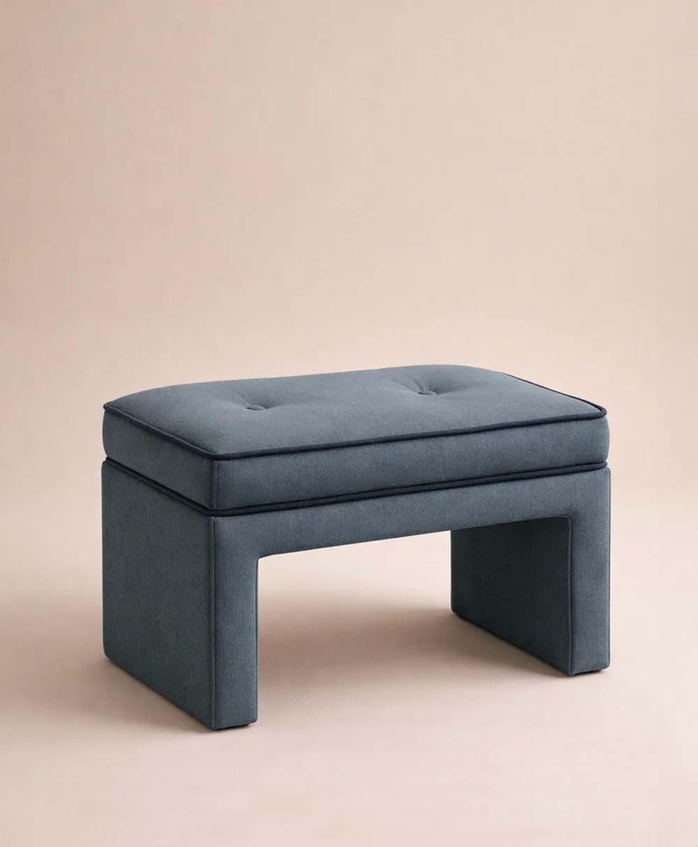 A modern, rectangular, teal upholstered ottoman with a cushioned top and open sides, set against a plain beige background.