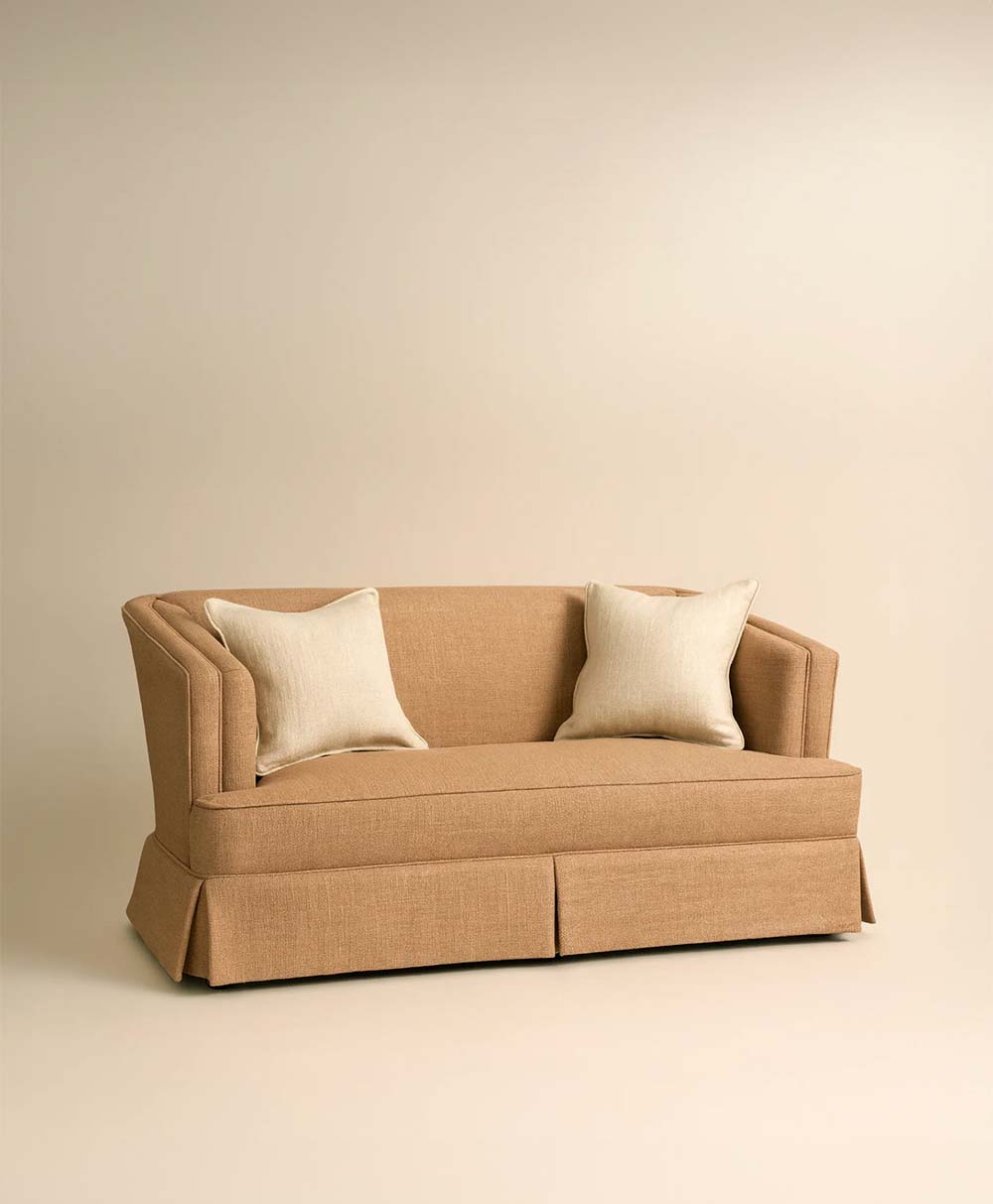 A tan upholstered sofa with a skirted base and two light beige throw pillows, set against a plain beige background.