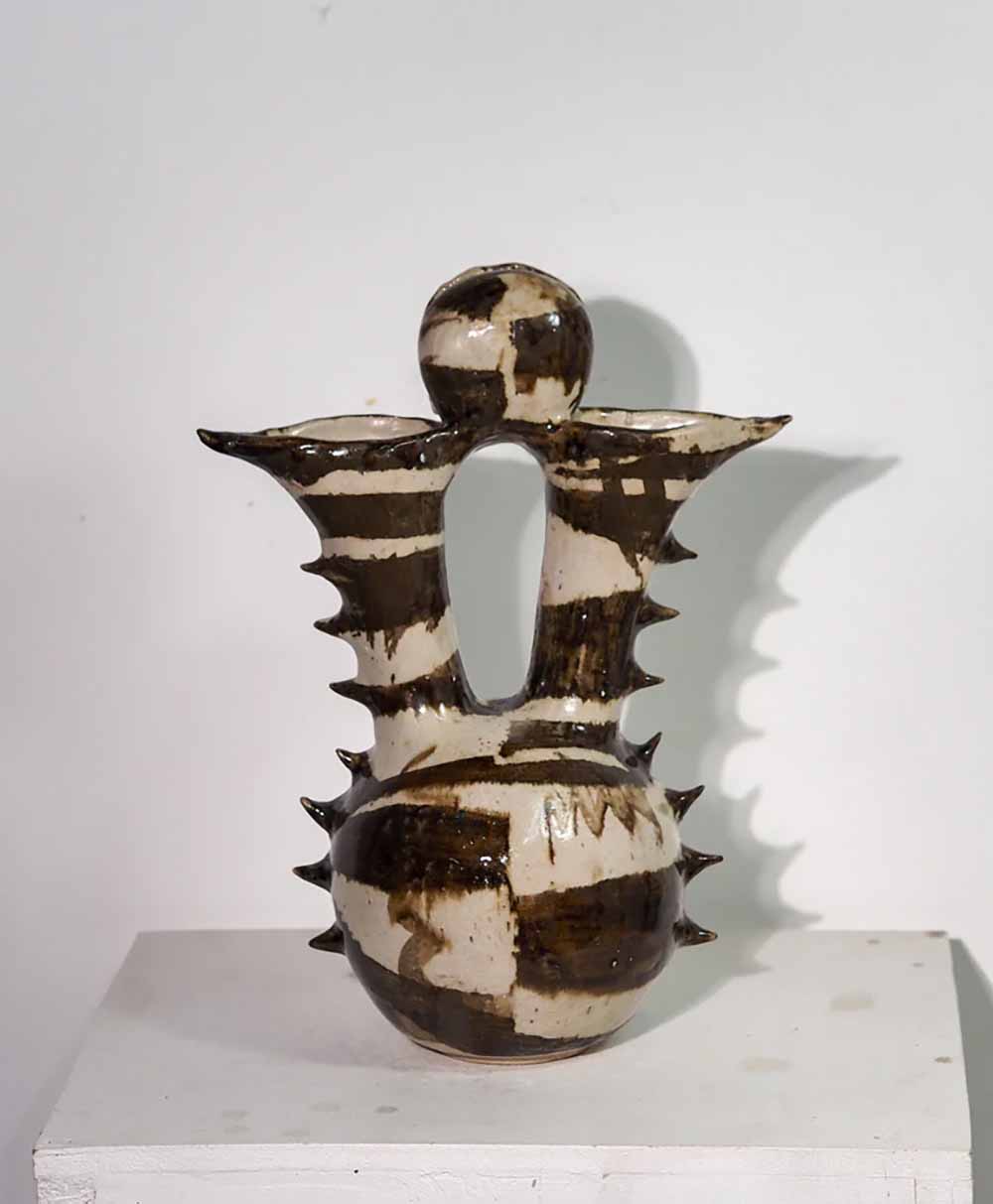 A ceramic vase with two spouted openings, a central sphere on top, and protruding spikes around the body. The surface features bold brown and white abstract stripes and patterns. The vase sits on a white pedestal against a plain wall.