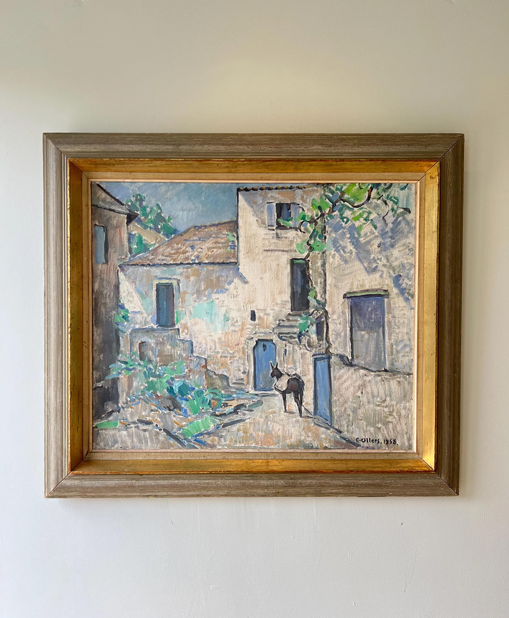 Framed painting of a sunlit Mediterranean village scene with old stone buildings, blue doors and windows, greenery, and a person standing in the courtyard. The artwork is signed and dated in the bottom right corner.