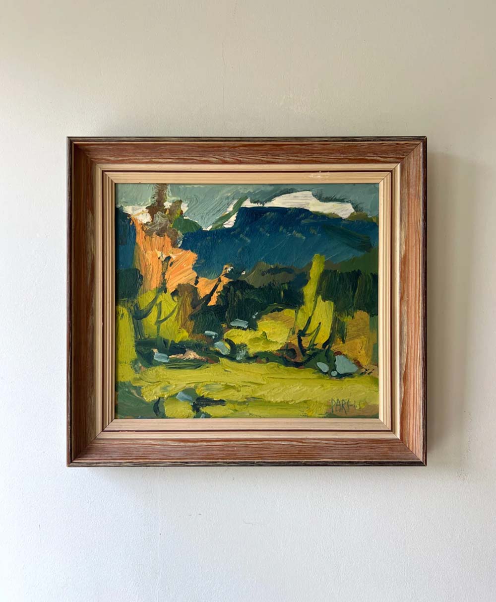 Framed painting of a colorful landscape with bold, broad brushstrokes, showing green fields, trees, and distant dark blue mountains beneath a light sky. The wall behind the frame is plain and light-colored.