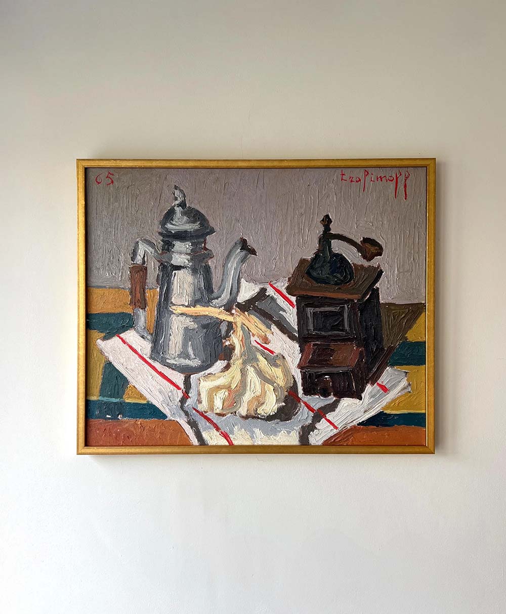 A framed painting of a still life featuring a silver coffee pot, a garlic bulb, a small hand-crank coffee grinder, and a striped cloth on a table, hung on a plain light-colored wall.
