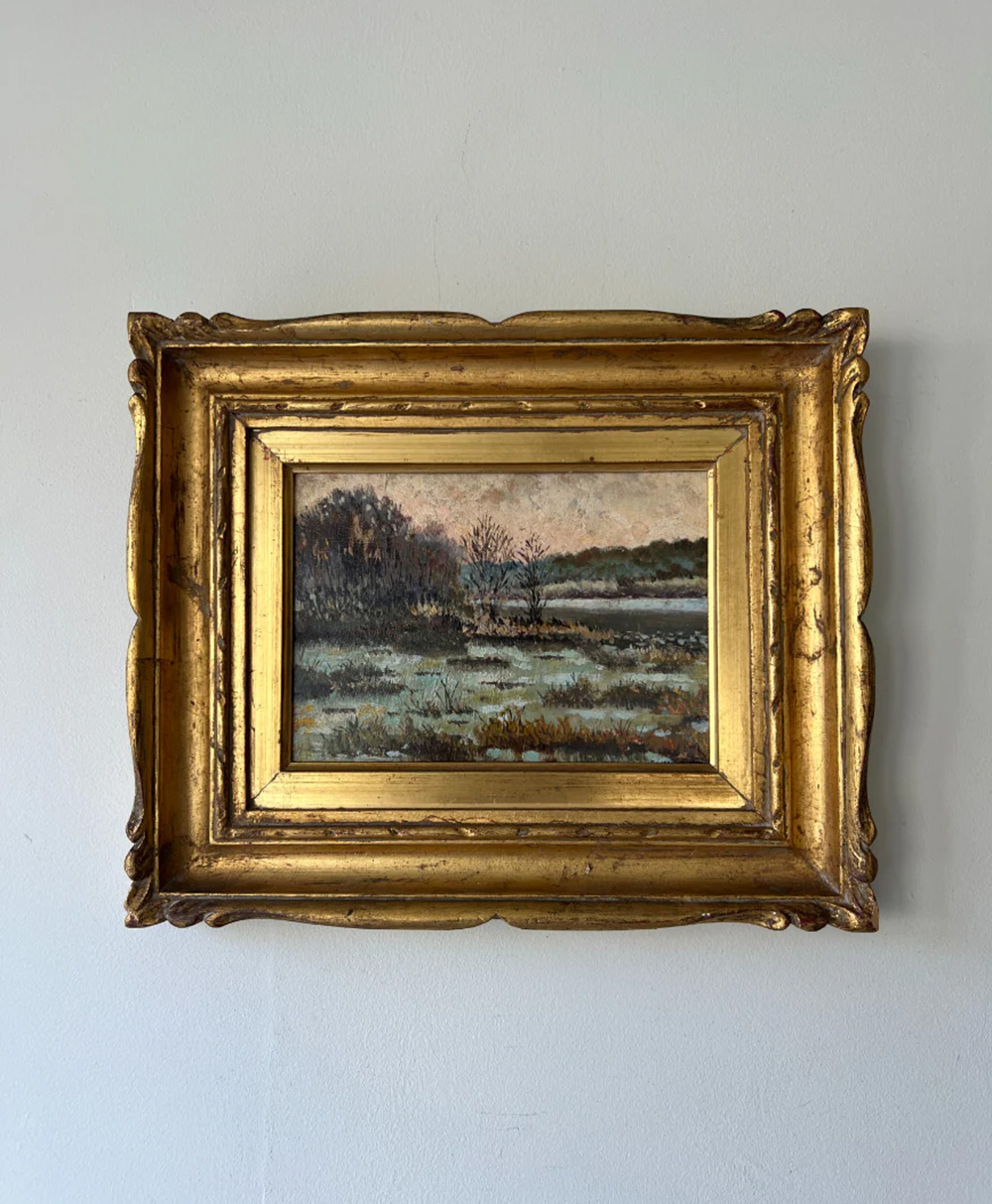 A gilded ornate frame hangs on a light gray wall, showcasing a landscape painting of a grassy marsh with trees and cloudy sky in muted green and brown tones.