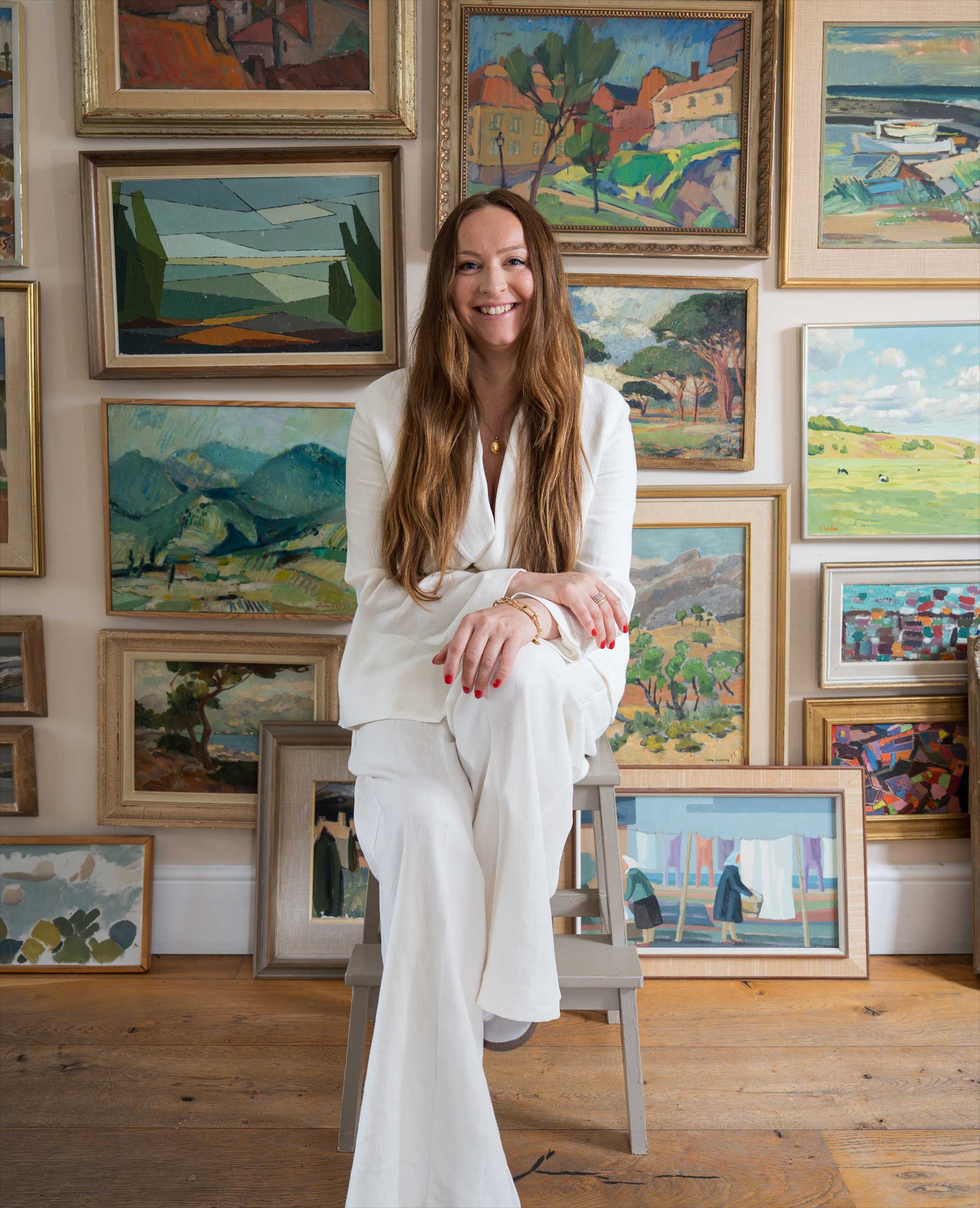 A woman with long brown hair, dressed in a white outfit, sits on a small step stool in front of a wall filled with colorful framed landscape paintings. The room has wooden floors.