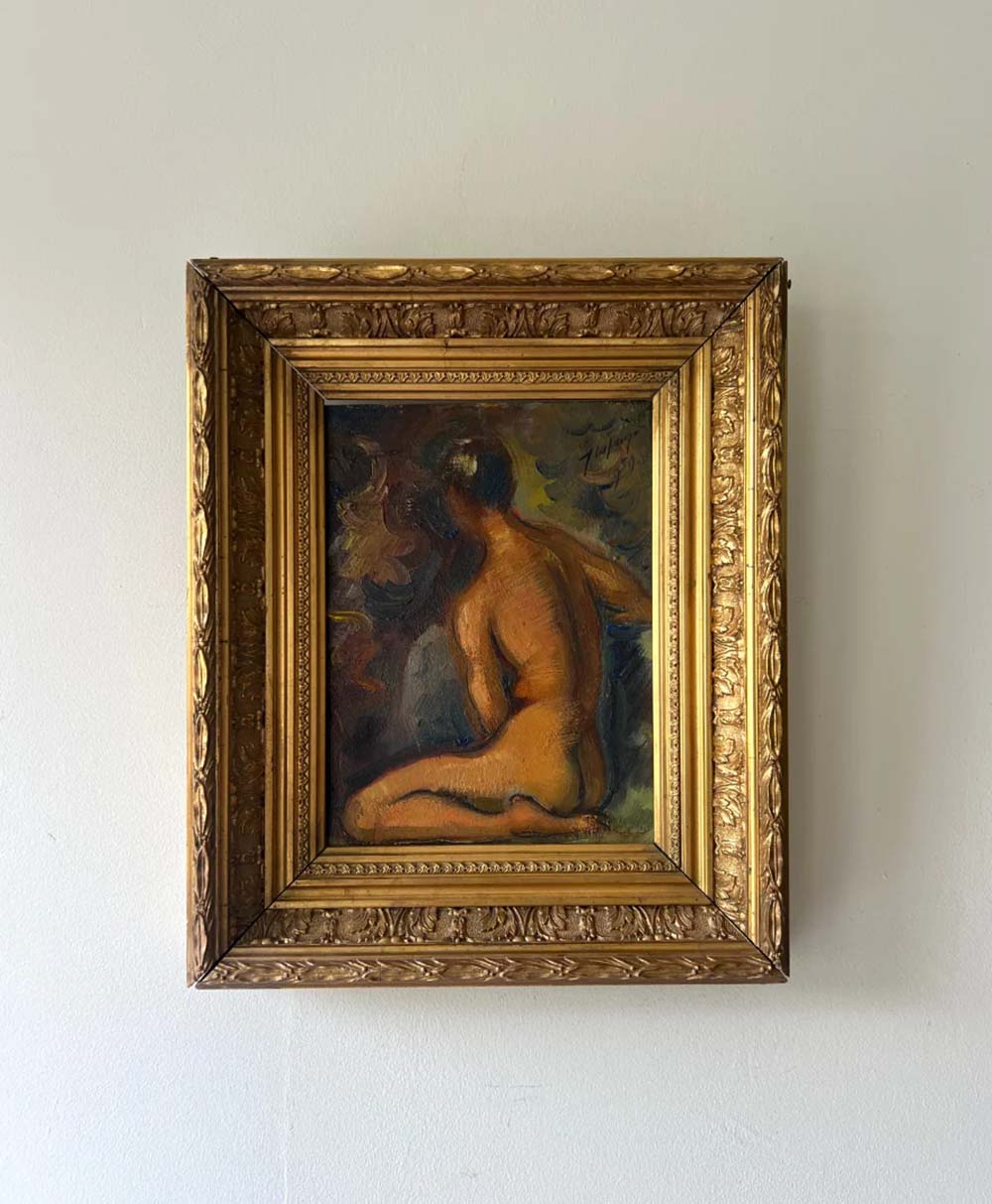 A framed painting of a nude figure sitting with their back turned, rendered in warm tones and set in an ornate gold frame, hangs on a plain white wall.