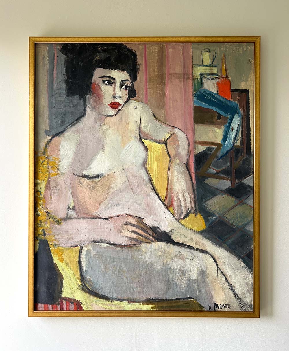 A framed painting of a seated nude woman with short dark hair, resting on a yellow chair against a background of abstract shapes and muted colors, with a table and bottles visible in the background.