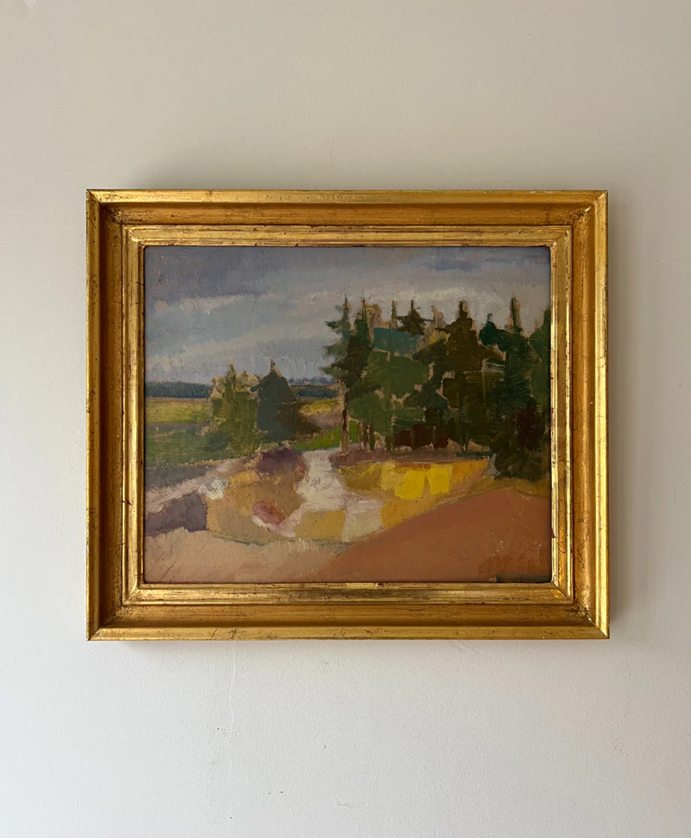A framed painting with a gold frame hangs on a plain light-colored wall, depicting a landscape with trees, a path, and patches of green, yellow, and brown under a blue sky.