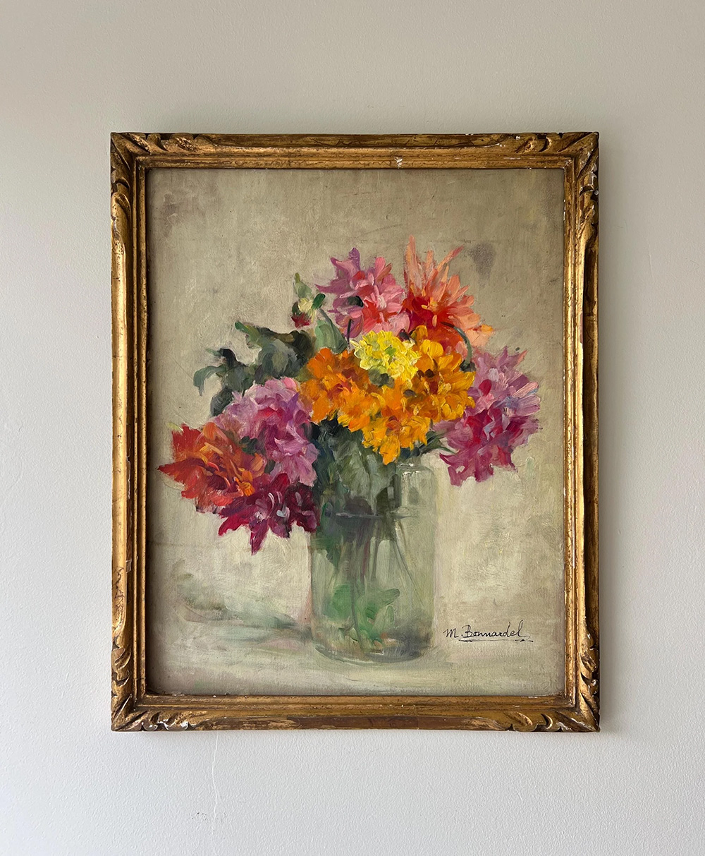 A framed painting of a bouquet of colorful flowers including yellow, orange, pink, and purple blooms in a glass jar, set against a plain light background and signed by the artist in the bottom right corner.
