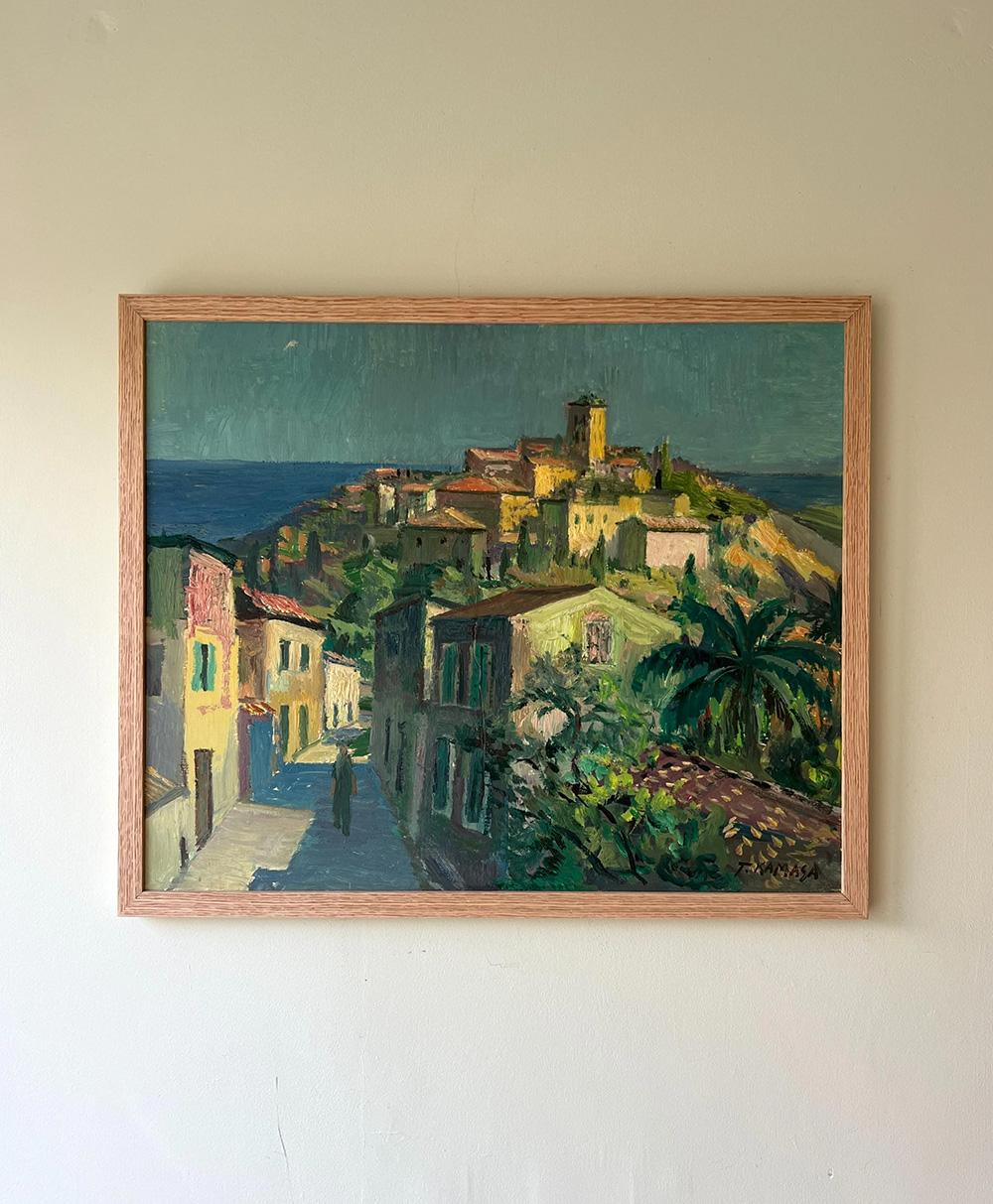 A framed painting of a coastal village with colorful houses, a winding street, palm trees, and a hilltop fort or castle under a blue-green sky, hanging on a plain light-colored wall.
