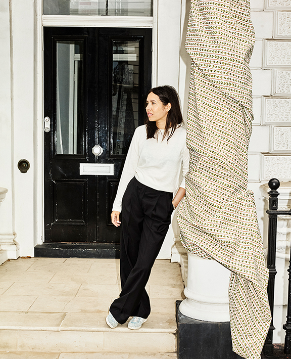 A woman in a white top and black pants stands on a stone porch in front of a black door, leaning against a white column wrapped in patterned fabric.