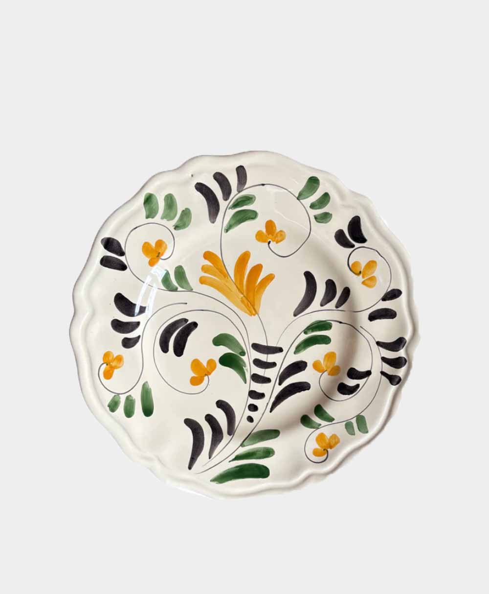 A round ceramic plate with a wavy edge, decorated with hand-painted green, black, and yellow floral patterns on a white background.