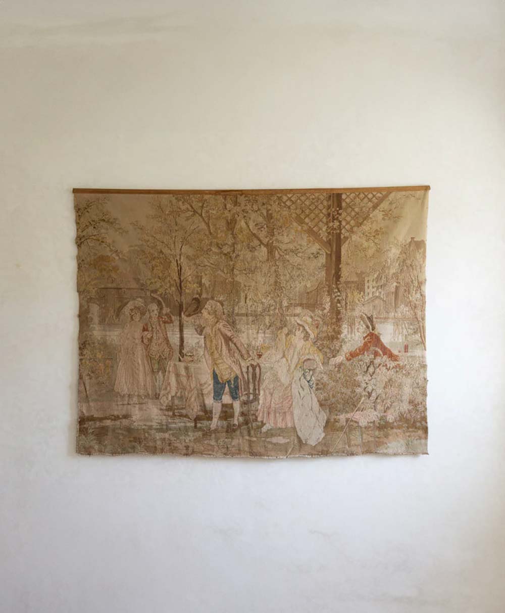 A faded tapestry hangs on a plain white wall, depicting an outdoor scene with several people in historical clothing standing and interacting in a garden or park setting.