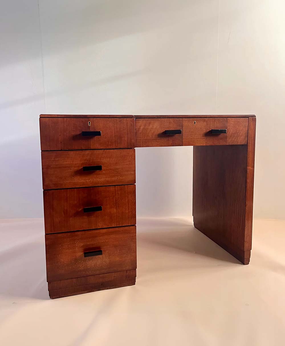 A vintage wooden desk with four drawers on the left and one wide central drawer, featuring black handles and a minimalist, mid-century design, set against a plain light background.
