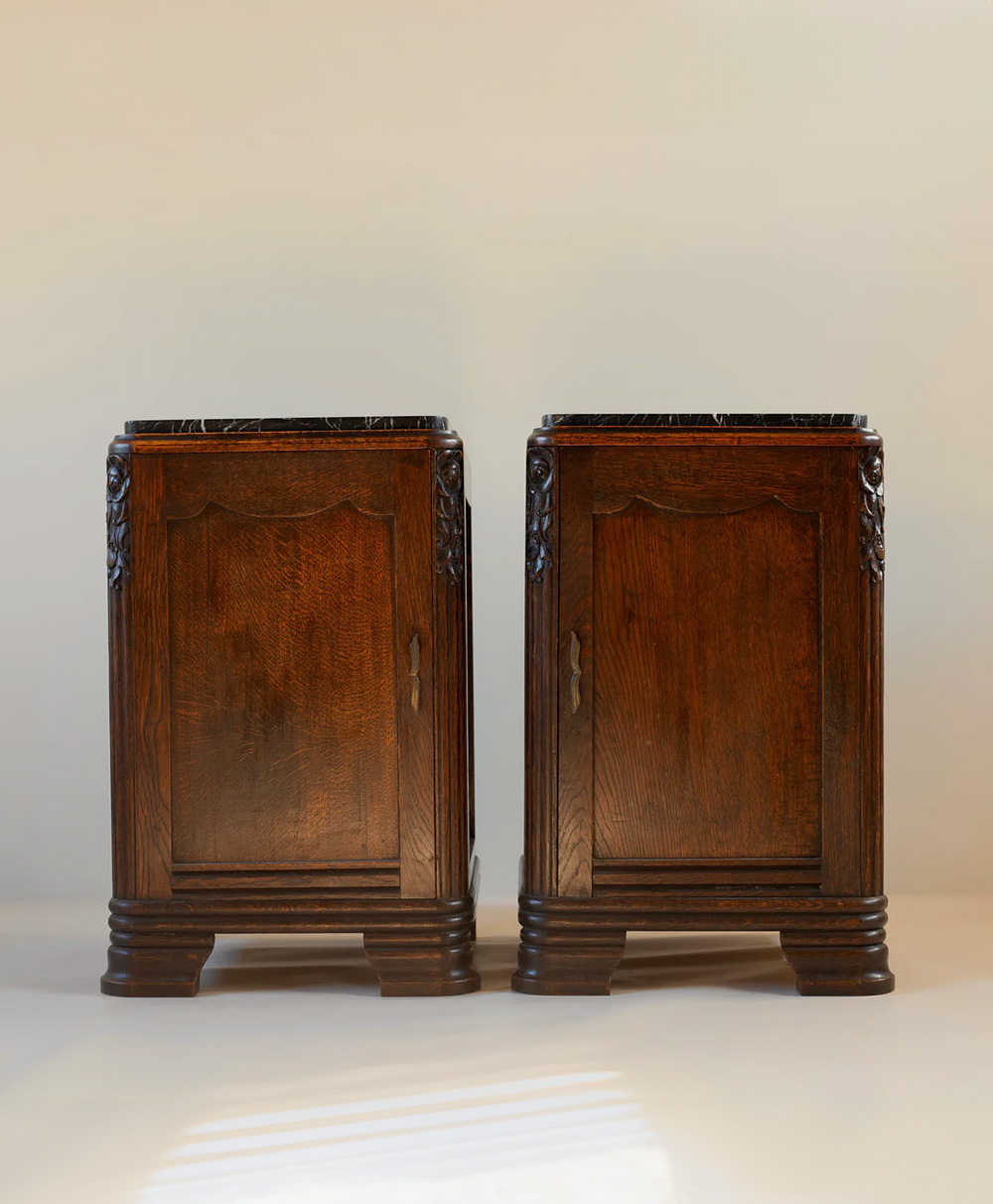 A pair of vintage wooden nightstands with carved details, paneled doors, and dark marble tops, standing side by side on a light background.