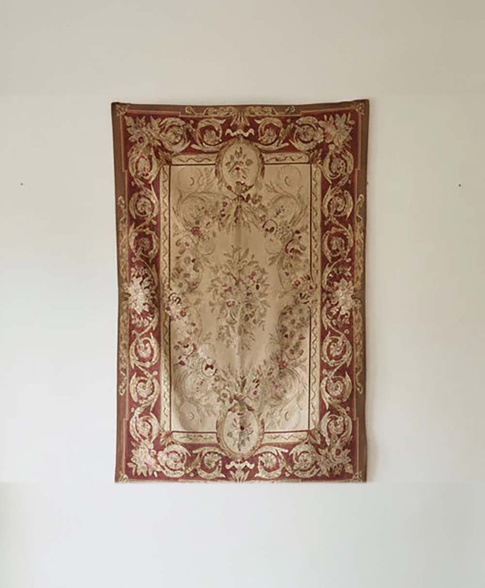 A decorative rectangular rug with ornate floral and vine patterns in shades of beige, red, and gold hangs on a plain white wall.