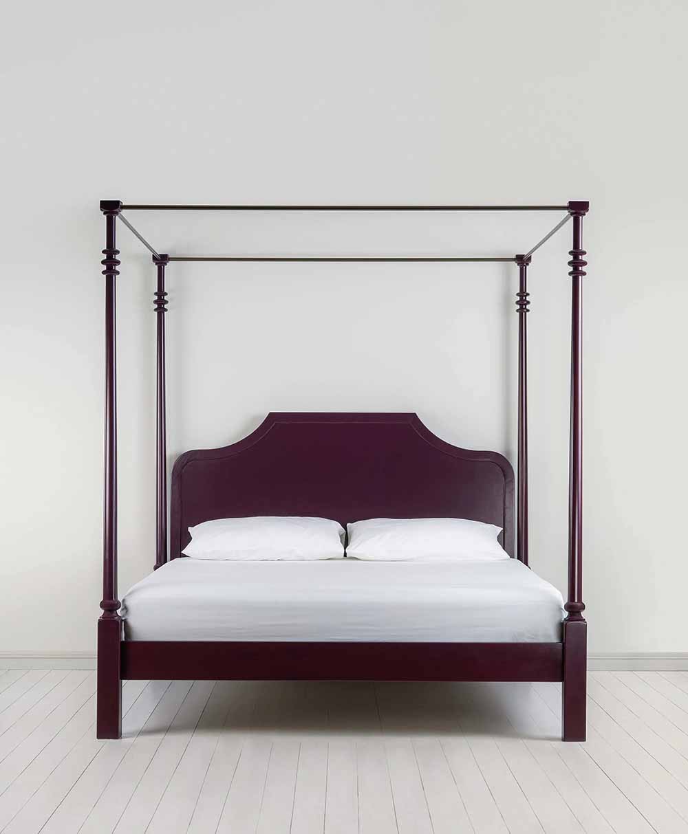 A dark wood four-poster bed with a high headboard, white bedding, and two pillows sits in a minimalist room with light-colored walls and pale wooden floorboards.