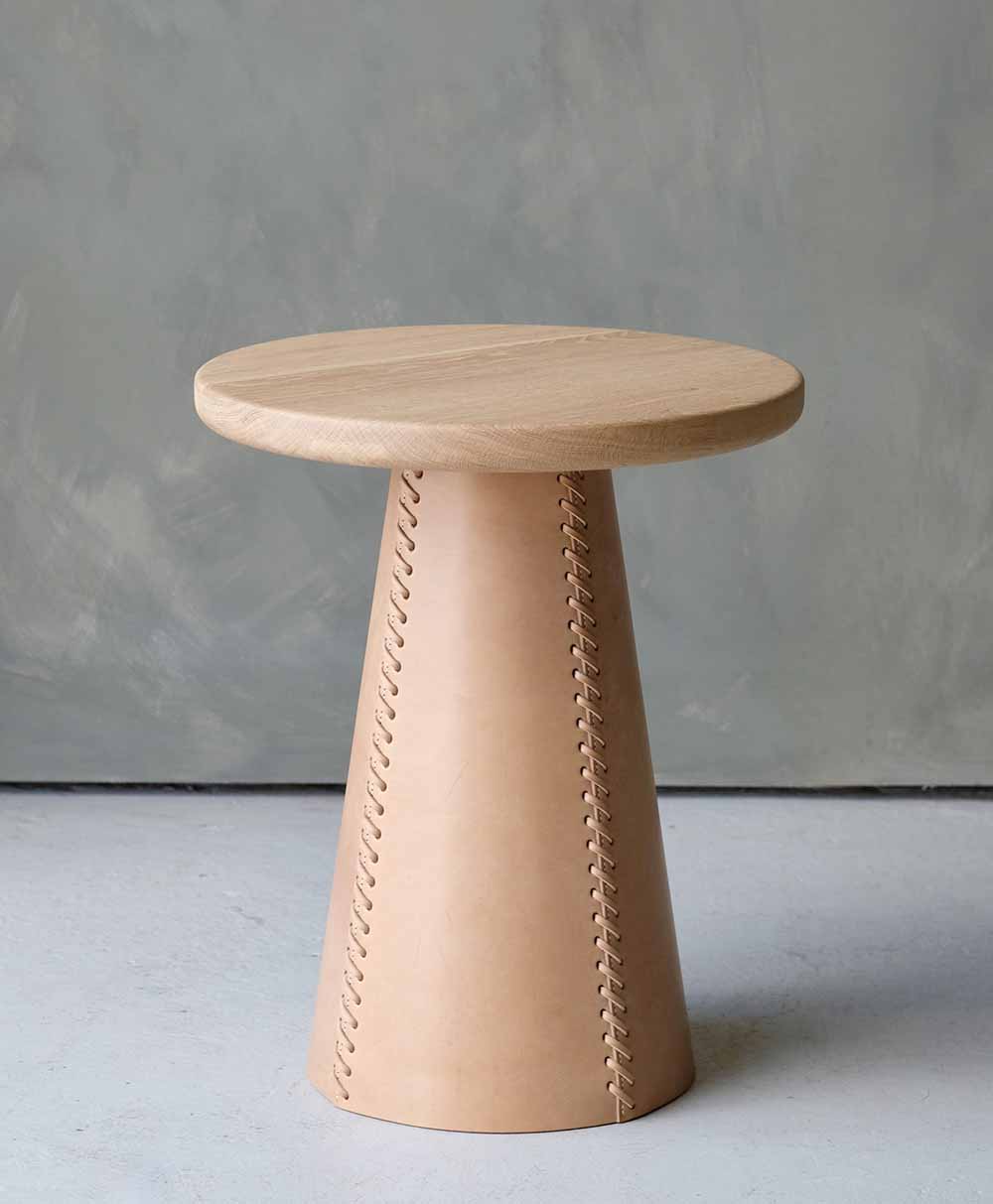 A round wooden stool with a smooth top and a conical base wrapped in light leather, featuring visible zigzag stitching along the front. The stool is set against a neutral gray background.