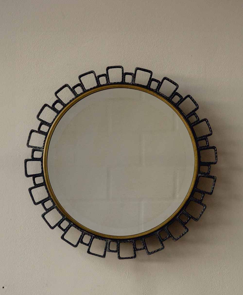 A round wall mirror with a decorative black metal frame featuring rectangular loops radiating outward, creating a sunburst effect, mounted on a plain beige wall.