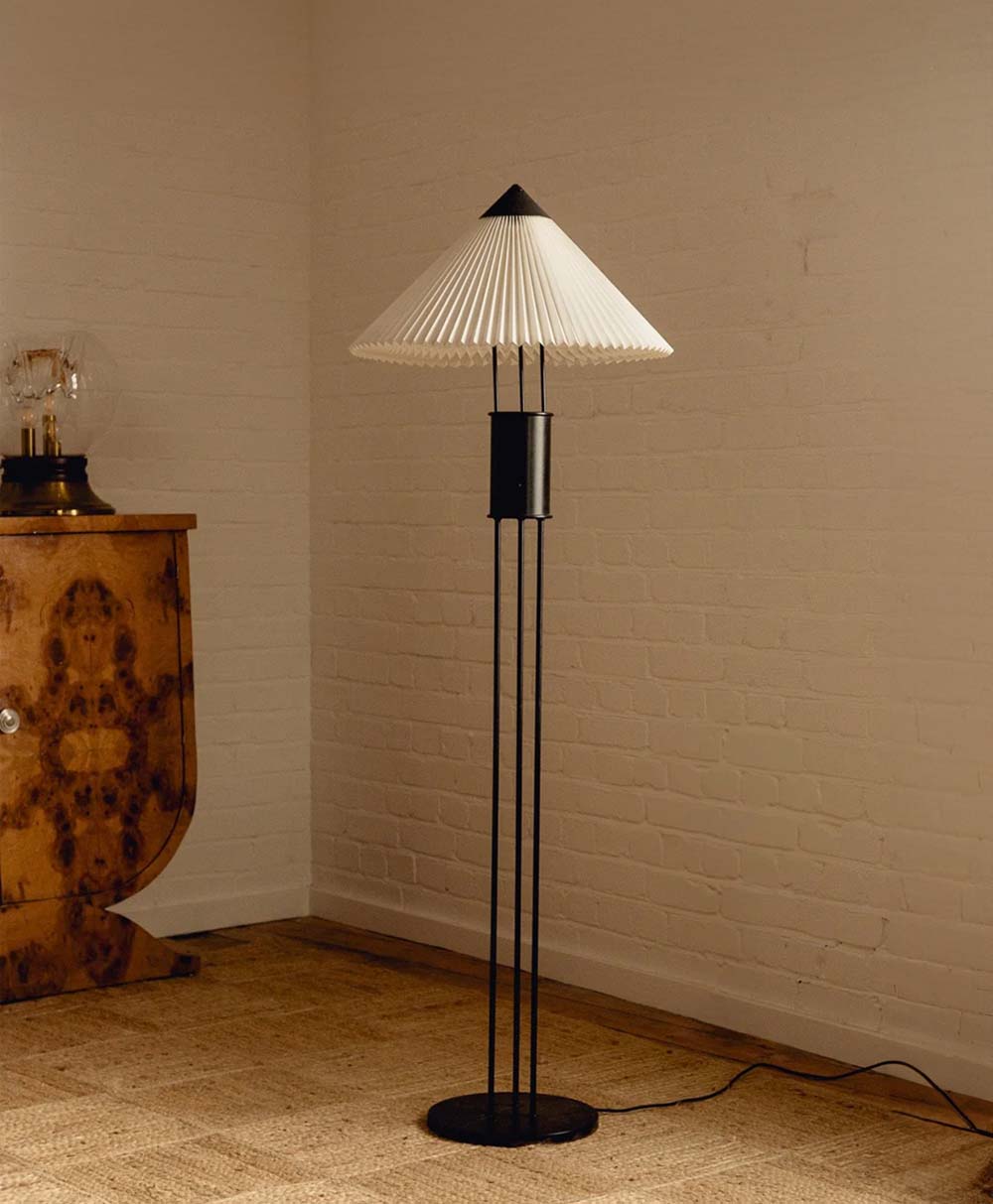 A modern floor lamp with a pleated white lampshade and black stand sits on a woven rug next to a wooden cabinet in a room with white brick walls.