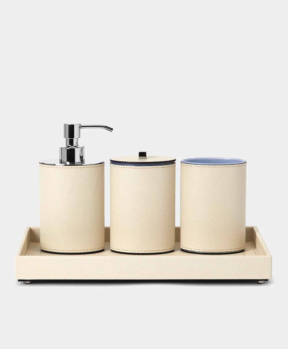 A beige bathroom set on a matching tray, featuring a soap dispenser, a lidded container, and an open cup, all with a textured finish and silver accents, against a plain light background.