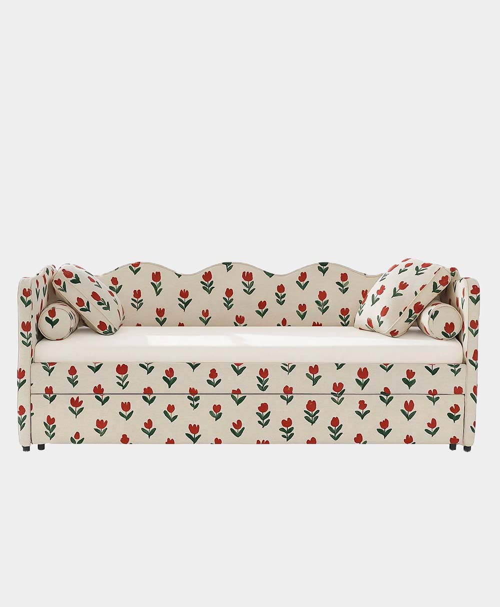 A cream-colored daybed with a wavy backrest, two matching pillows, and a red and green tulip floral pattern covering the entire surface, set against a plain white background.