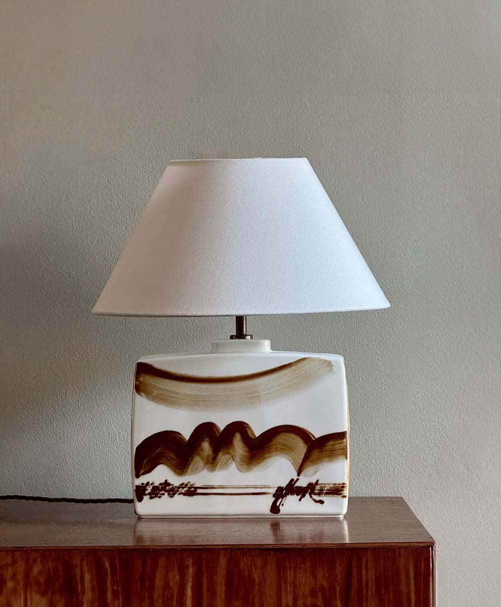 A table lamp with a white rectangular ceramic base featuring brown abstract brushstrokes, topped with a wide white lampshade, sits on a wooden surface against a light gray wall.