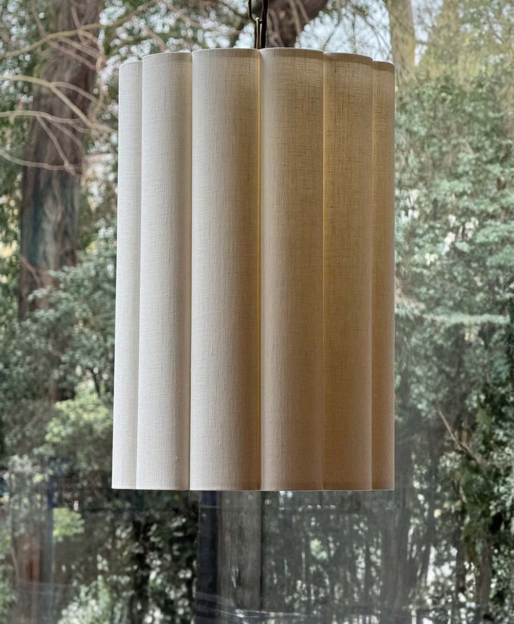 A large, cylindrical, pleated beige lampshade hangs indoors in front of a window, with trees and greenery visible outside in the background.
