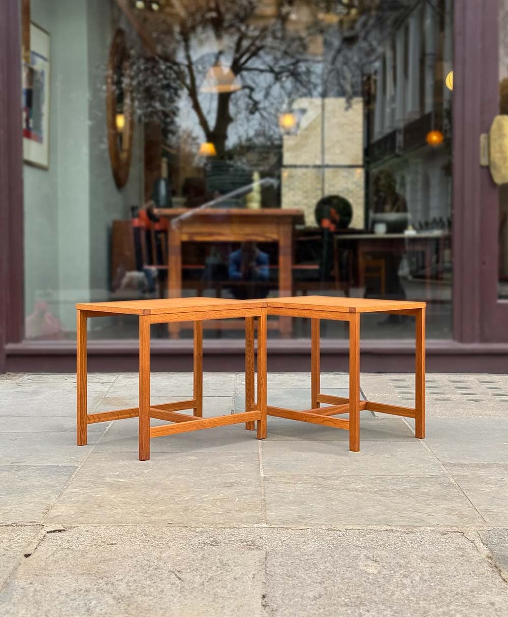 Two wooden tables with angular legs are positioned together on a stone pavement in front of a large glass window, reflecting trees and warm lights from inside a building.
