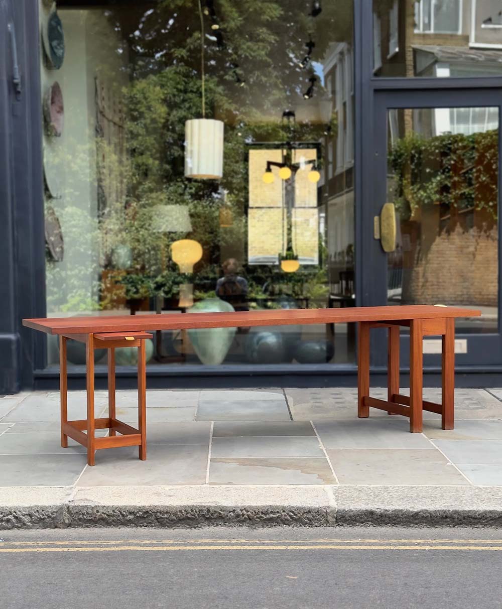 A wooden table and small side table are displayed on a sidewalk in front of a glass storefront with reflections of hanging lights, trees, and pottery inside.