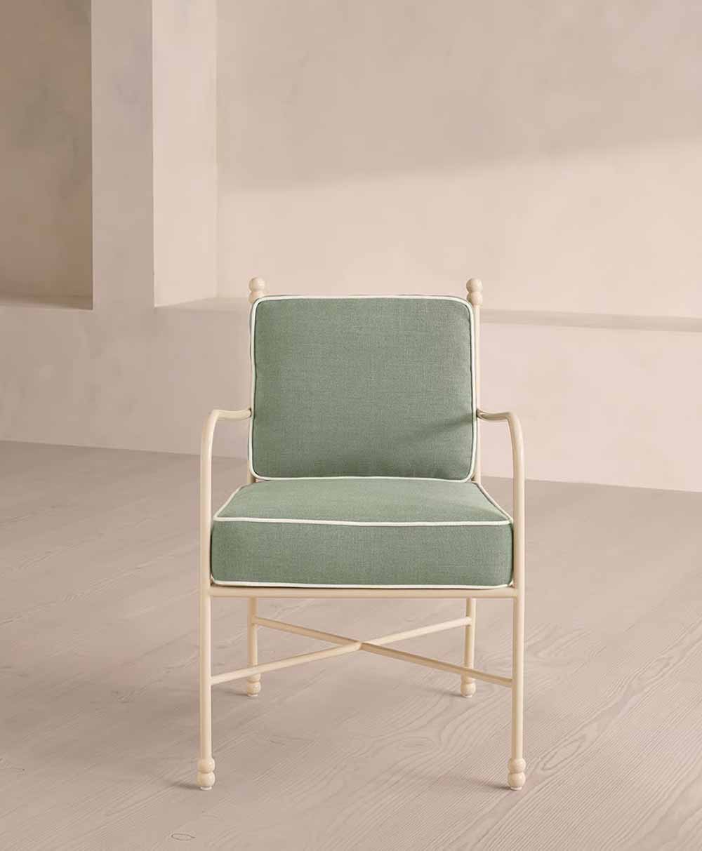 A light green cushioned chair with white piping and a cream-colored metal frame, placed on a light wood floor against a beige wall in a minimal, modern setting.