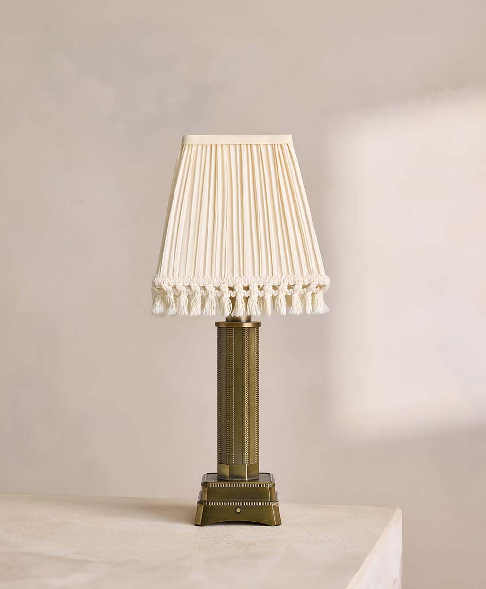 A vintage-style table lamp with a pleated, cream-colored fabric lampshade and tassel trim, standing on a rectangular, ornate brass base, placed on a light surface against a neutral background.