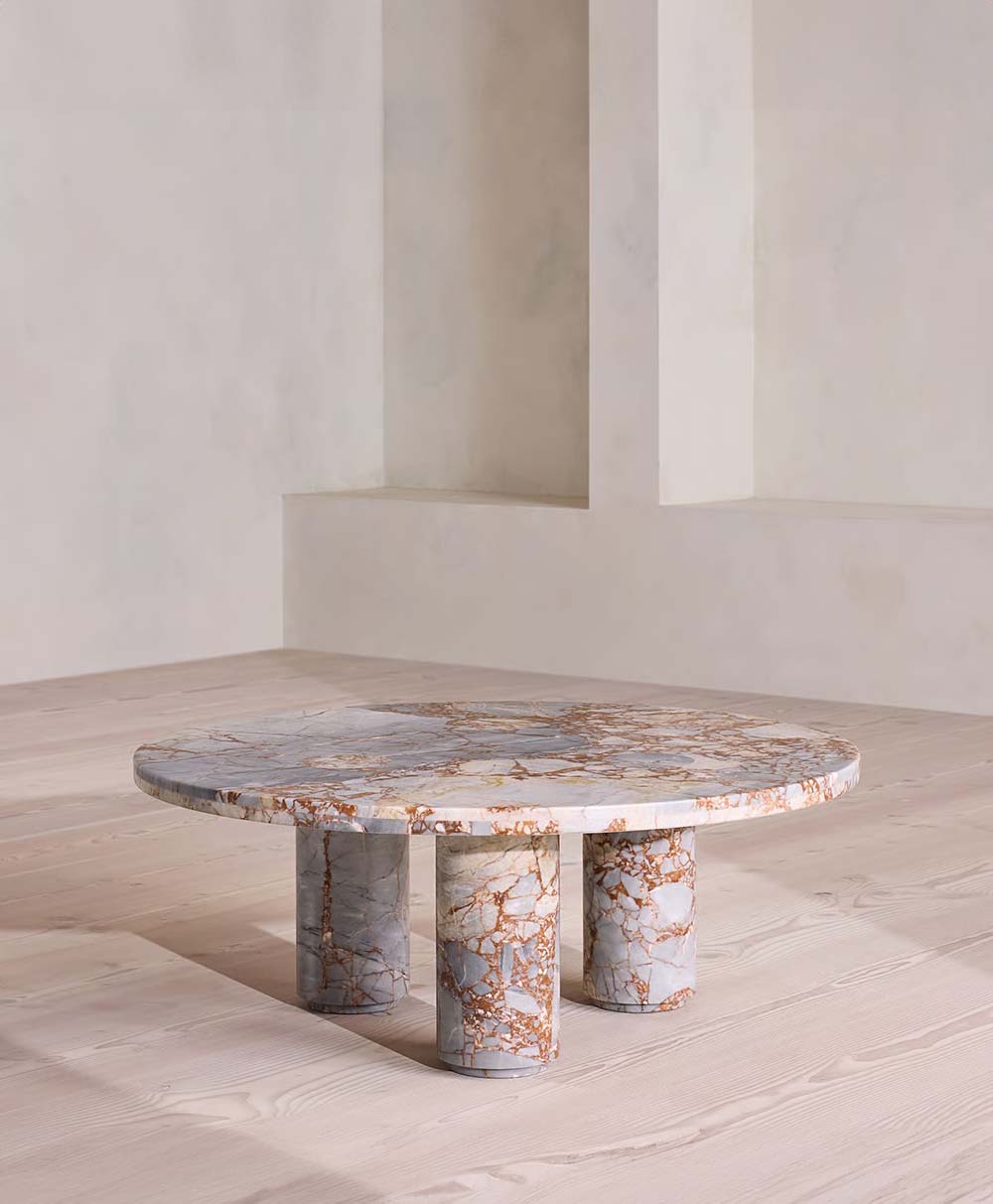 A round marble coffee table with a white and brown veined surface, supported by three thick cylindrical legs, sits on a light wood floor in a minimalist, light-toned room.
