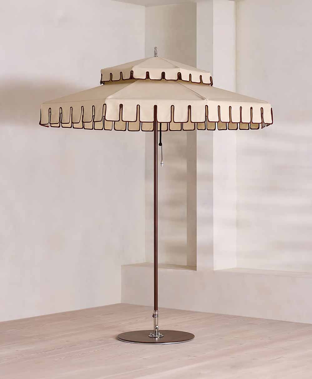 A beige outdoor umbrella with brown trim and a scalloped edge stands on a round metal base in a minimal, light-colored indoor space with wooden flooring and plain walls.