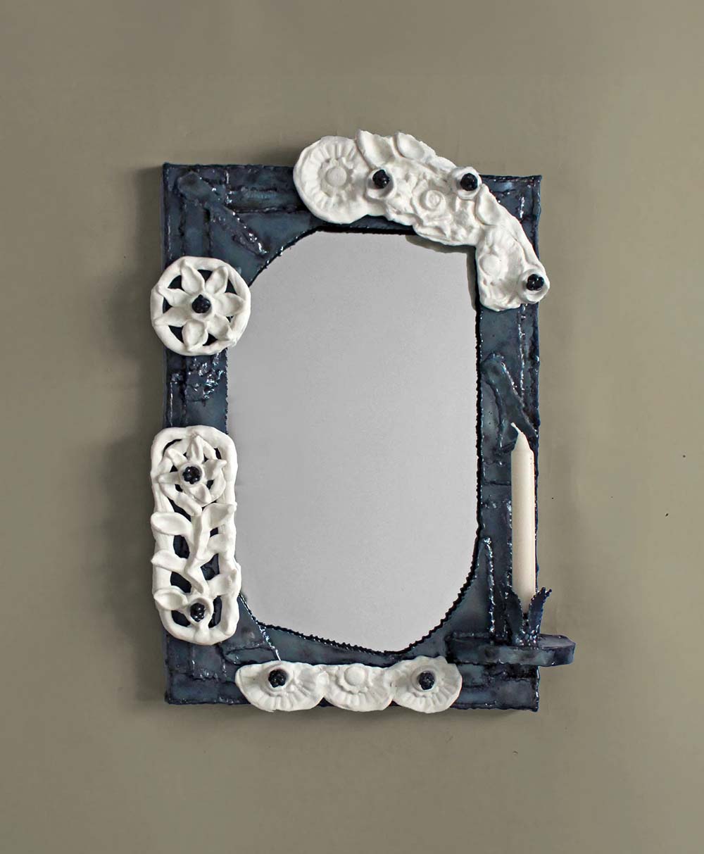 A rectangular mirror with a dark blue frame decorated with white ceramic floral and shell designs. A white candle is held by a small holder attached to the right side of the frame. The background is a plain beige wall.