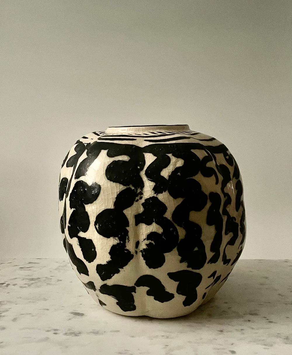 A round ceramic vase with a cream-colored base and irregular black abstract patterns displayed on a marble surface against a plain light background.