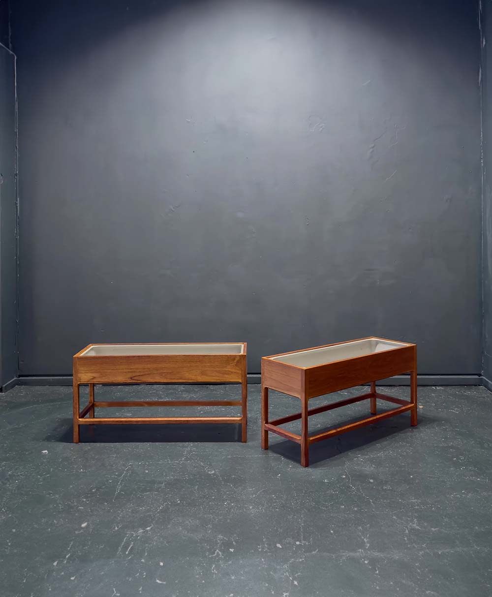 Two rectangular wooden tables with glass tops stand side by side on a dark, slightly worn floor against a plain, dark gray wall in a minimalist setting.