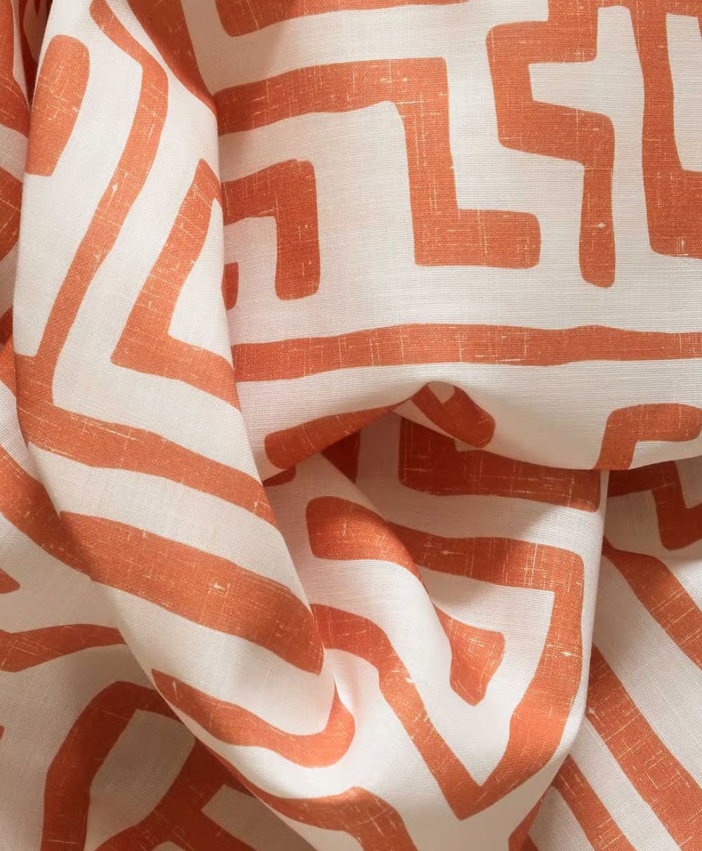 A close-up of white fabric with an abstract, maze-like pattern in a terracotta orange color. The cloth is loosely draped, creating soft folds and shadows.