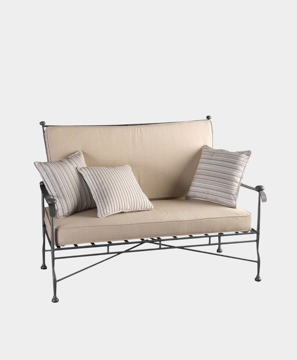 A beige cushioned metal loveseat with three striped throw pillows, featuring black metal frame and armrests, set against a plain white background.