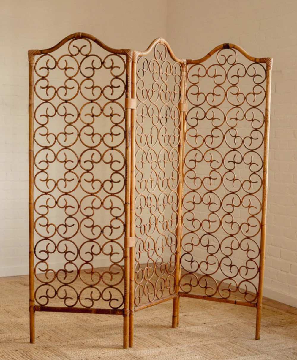 A decorative three-panel rattan room divider featuring intricate swirling patterns stands on a woven rug in a light-colored room with white brick walls.