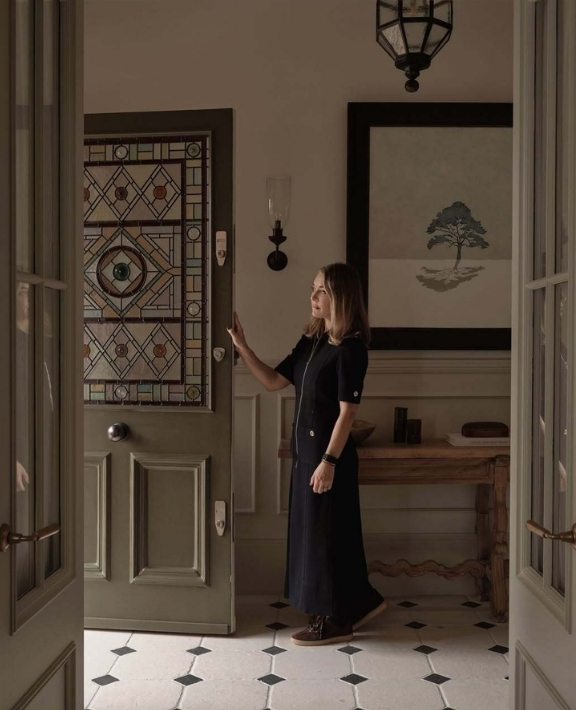 A woman in a long black dress stands inside a room, reaching for the handle of a wooden door with a colorful stained glass window. The room has a patterned tile floor, a large framed tree artwork, and a wooden table.