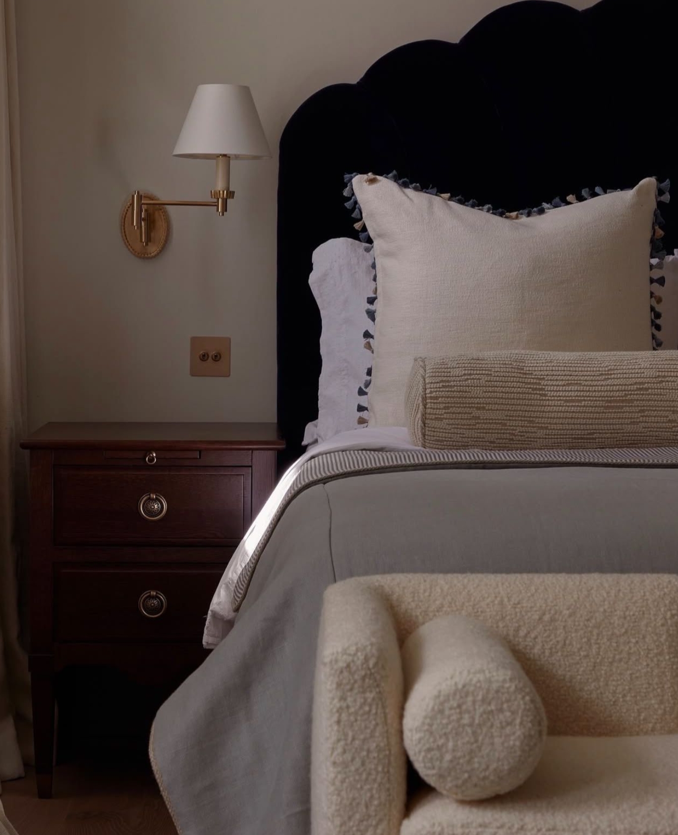 A cozy bedroom corner with a dark upholstered headboard, white pillows, a beige bolster, a gray blanket, a wooden nightstand with drawers, a wall-mounted lamp, and a cream-colored textured armchair in the foreground.