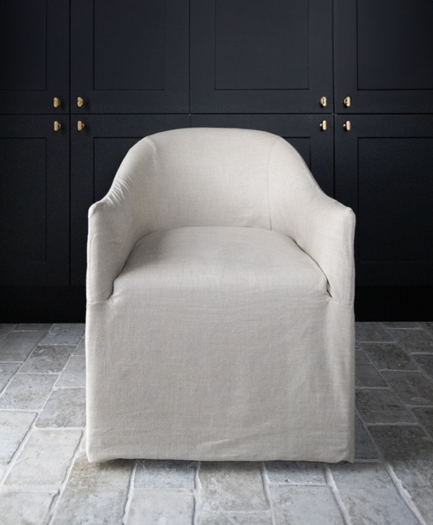 A light beige, upholstered armchair with a skirted base sits on a stone tile floor in front of black paneled cabinets with brass hardware.